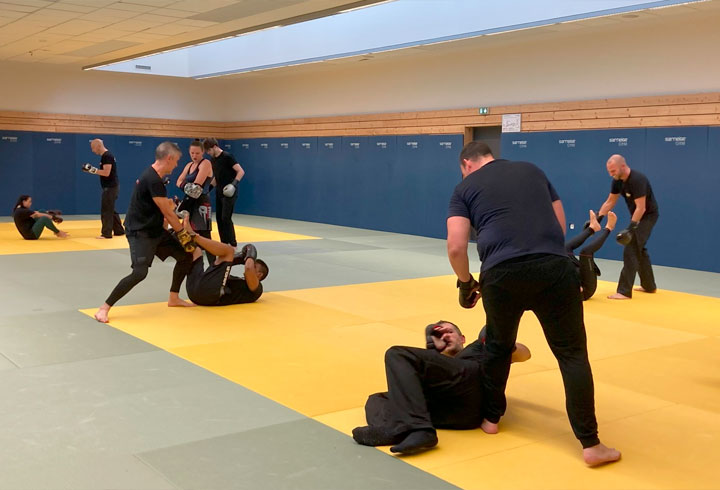 Licenciés du Dojo 77 en pleine séance d'entraînement de MMA, effectuant des techniques au sol avec concentration sous la supervision d'un instructeur.
